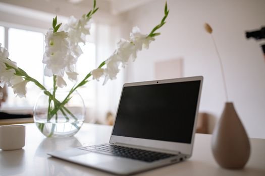 A minimalist workspace with a silver laptop and white flowers in a vase on a table.